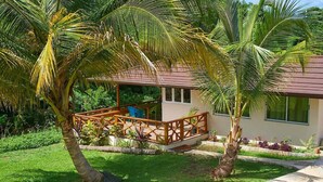 Un espacio al aire libre cubierto con una terraza de madera, rodeado de palmeras y un césped cuidado.