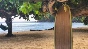 Escena de playa con una tabla de surf bajo un árbol, arena y mar tranquilo.