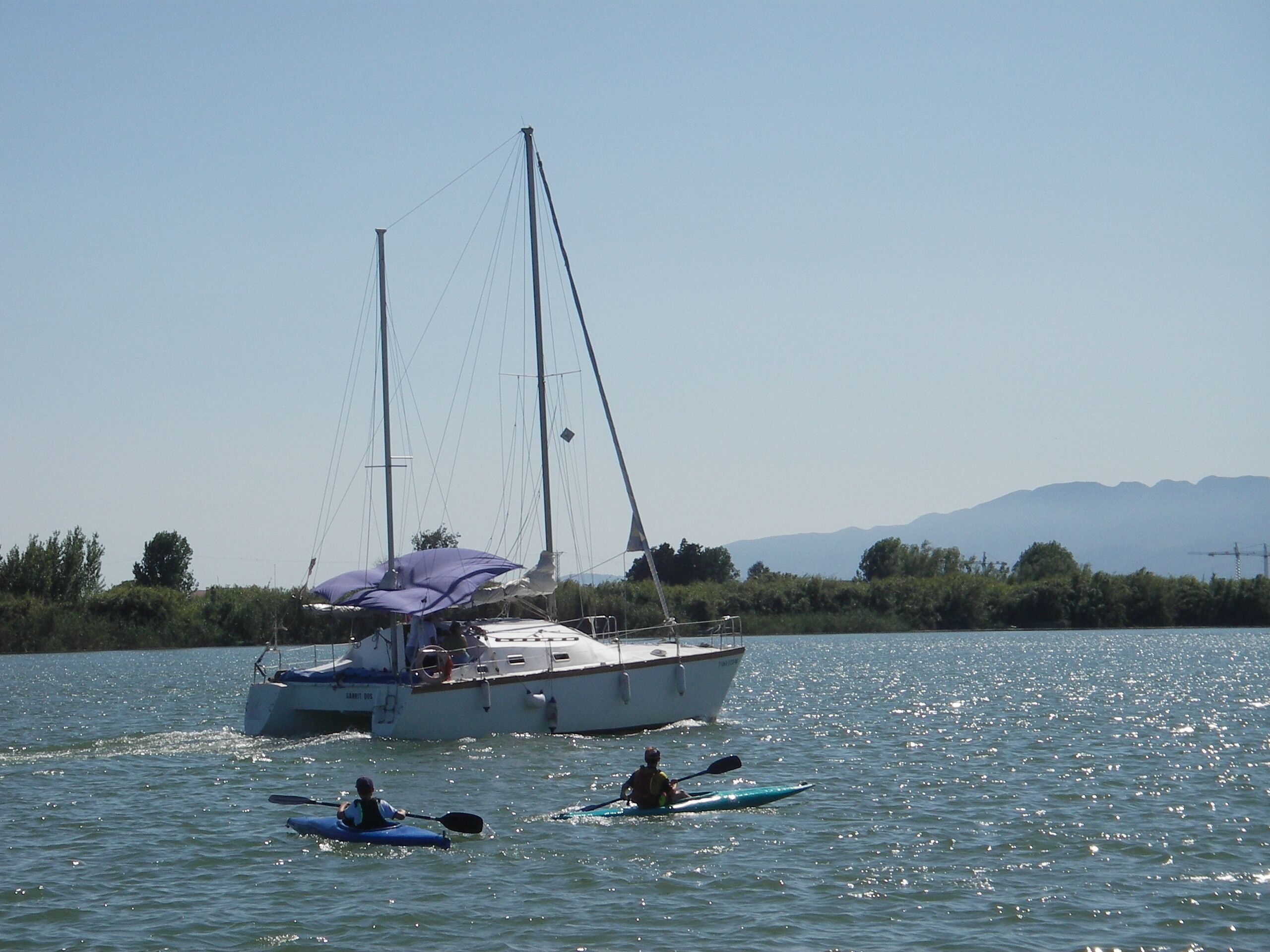 A sailboat with a canopy on the water, a person paddling a kayak, and a mountainous landscape in the background.