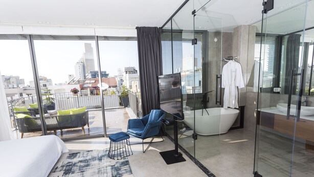 Ein modernes Hotelzimmer mit einem großen Bett, einer Sitzecke mit Sessel und Hocker, einem Glaskastenbad mit Badewanne und Blick auf die Stadt durch große Fenster.