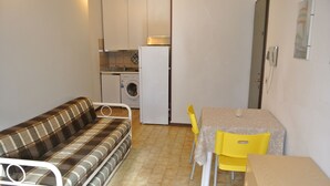 A compact living space with a sofa, a dining table with chairs, a washing machine, and a refrigerator. - Schubert 1 - Lignano (Lignano Sabbiadoro)
