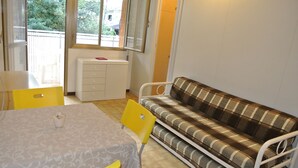 A room with a dining table, two yellow chairs, a plaid sofa, and a window with a view of greenery outside. - Schubert 1 - Lignano (Lignano Sabbiadoro)