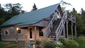 A rustic wooden house with a metal roof, a wooden staircase leading to a deck, and a garden with various plants and flowers.