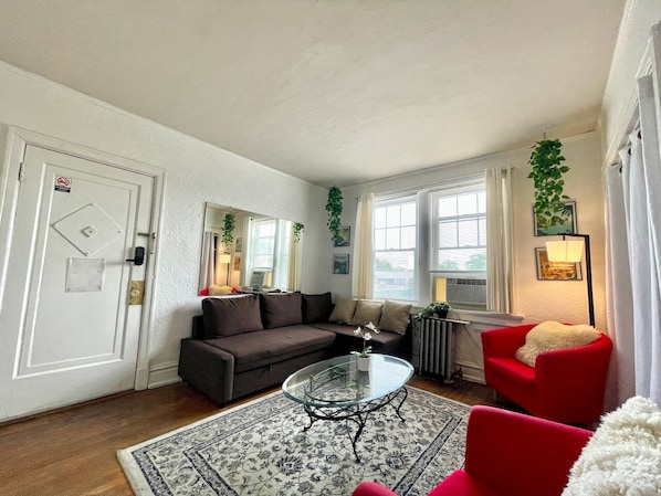 A living room with a dark brown sofa, a glass coffee table, a red armchair, and a white armchair. There is a door with a no smoking sign, a radiator, and a plant on the wall.
