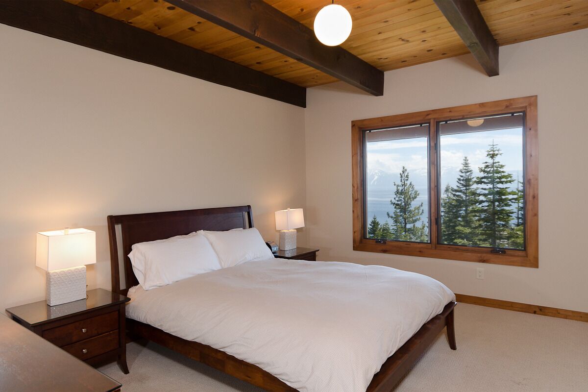 A bedroom with a wooden bed, two bedside tables with lamps, and a large window with a mountain view.