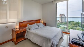 Un dormitorio con un ventanal grande, una cama con cabecera de madera, una mesita de noche y un escritorio con una computadora y controles remotos.