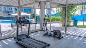 Un gimnasio con cintas de correr y una máquina elíptica, ventanas amplias y vistas a una piscina y edificios.