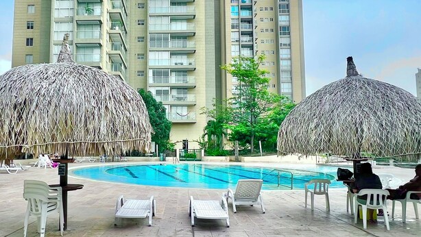 Zona de piscina con sillones de descanso y sombrillas de paja, rodeada de edificios residenciales altos.