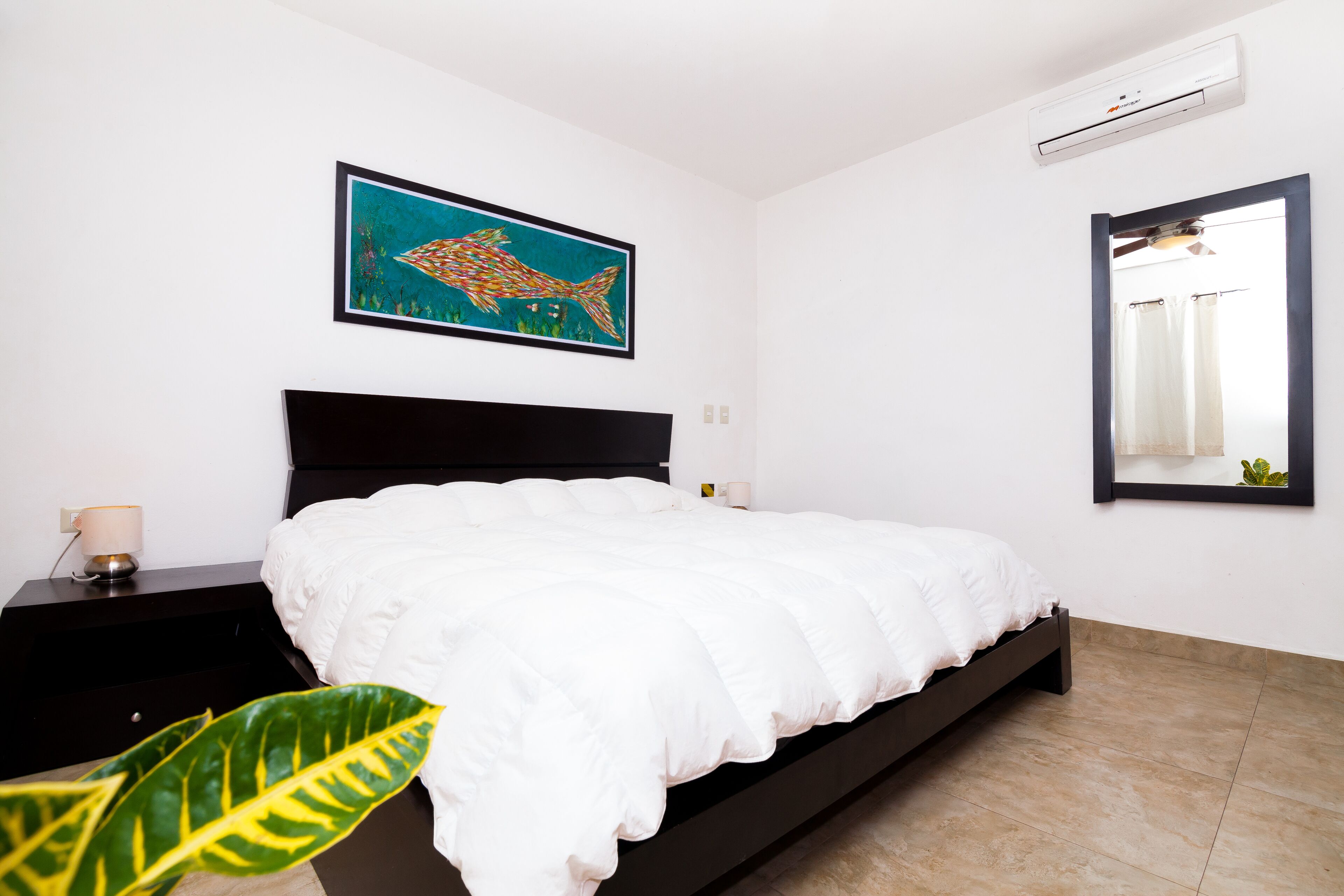 A bedroom with a bed, a nightstand, a wall-mounted air conditioner, a framed fish painting, and a mirror.