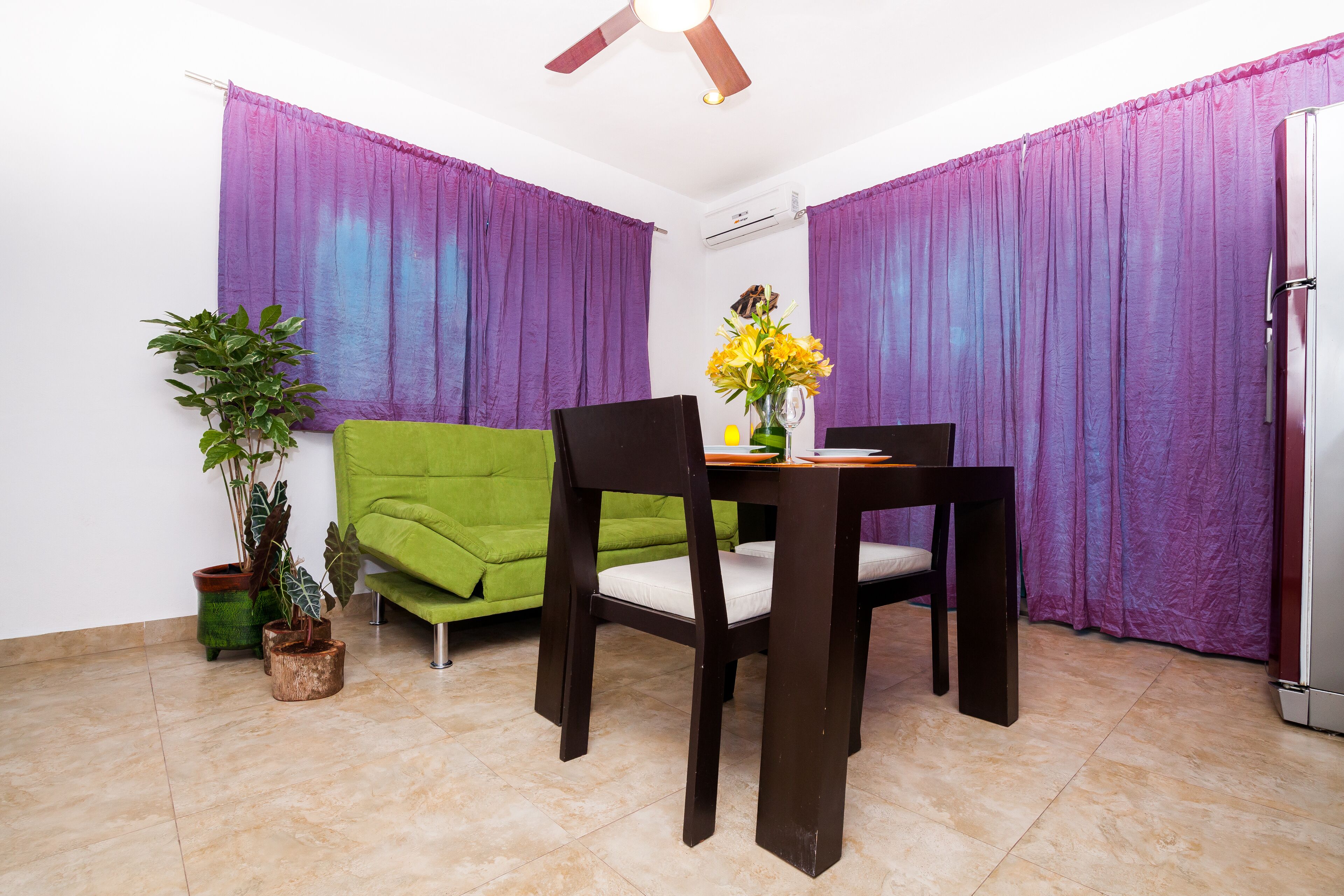 A dining area with a table, chairs, a green sofa, purple curtains, a ceiling fan, and a refrigerator.