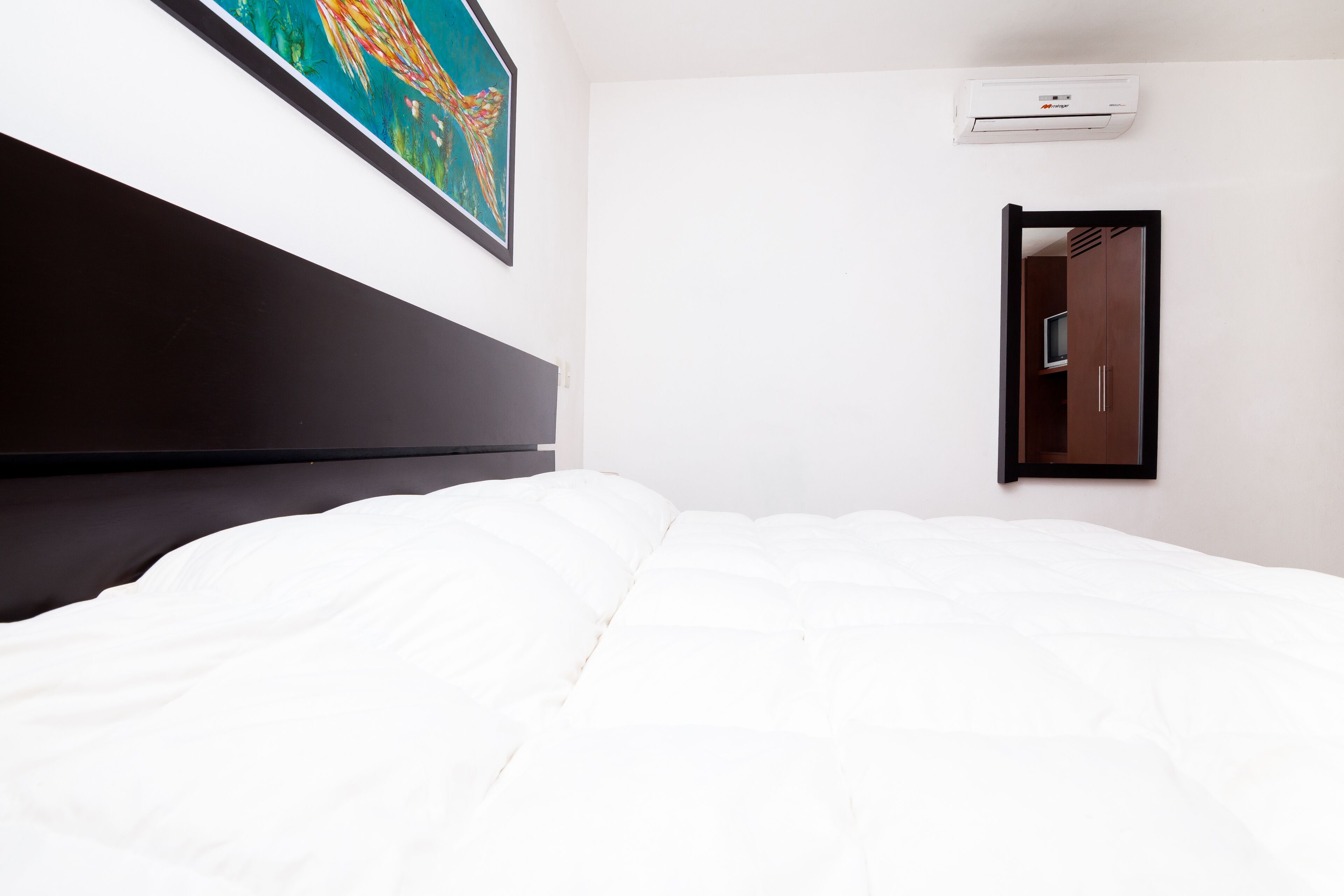Une chambre d’hôtel avec un grand lit, une tête de lit, un climatiseur fixé au mur, un tableau encadré et une porte dotée d’un miroir.