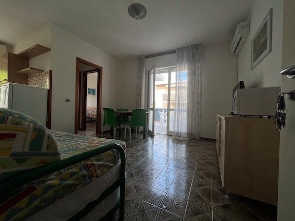A room with a bed, a kitchenette, a dining area with chairs, and a view through a door.