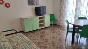 Une pièce avec un canapé vert, un téléviseur posé sur un meuble, une table à manger avec des chaises et des carreaux de sol à motifs.