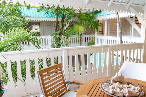 Una silla y una mesa de madera en un balcón con una cerca blanca de piquetes, rodeado de abundante vegetación y vistas a una piscina.