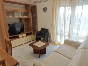A living room with a TV, wooden entertainment unit, a sofa, and a chair. - Condominio Billa 2 - Lignano (Lignano Sabbiadoro)