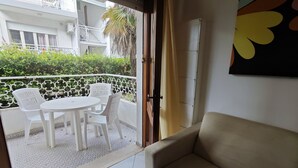 A balcony with a white table and chairs, a beige sofa, and a view of a building with balconies and greenery.
