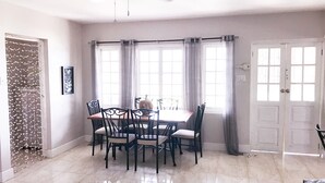 A dining area with a table and chairs, a window with curtains, a door, and a wall with a decorative panel. - YD Rooftop Lounge ocean view salt water pool with mountain view large 2bdrm  (Montego Bay)