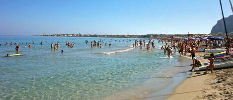 A crowded beach with clear water, people swimming and sunbathing, and boats on the shore.