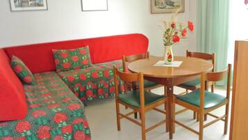 Ein Zimmer mit einem roten Sofa, einem grün gemusterten Sofa, einem Holzesstisch mit grünen Stühlen und einer Vase mit Blumen.