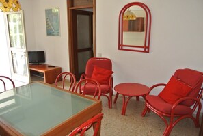 Une pièce avec une table à plateau de verre, des chaises rouges, une petite table ronde, un téléviseur posé sur un meuble en bois et un miroir accroché au mur.
