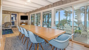 A modern dining area with a wooden table and blue chairs, a television, and a view of the sea through large glass doors.