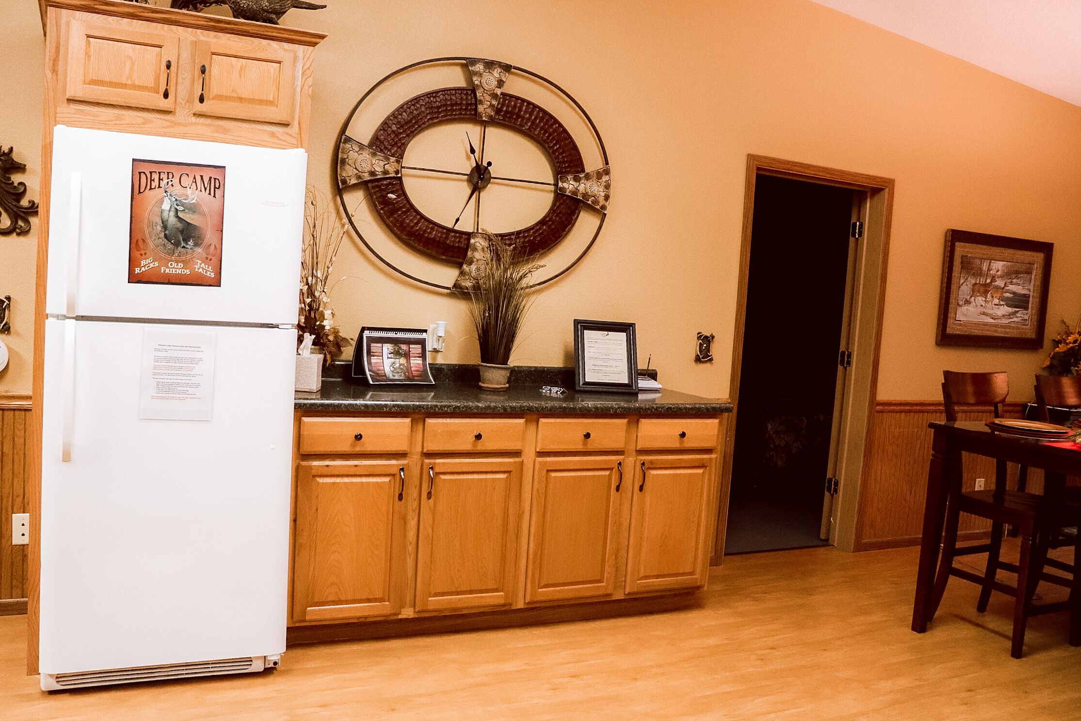 A kitchen with wooden cabinets, a refrigerator, a wall clock, and a dining area with chairs.