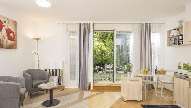 A modern living room with a sofa, a coffee table, and a dining area with a table and chairs. There is a kitchen area with cabinets and a window overlooking a garden.