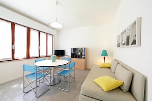 Un salon moderne avec un canapé beige, une table de salle à manger ronde entourée de chaises bleues, un téléviseur et un meuble avec des portes en verre.