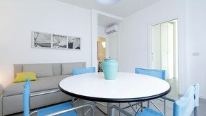 A dining area with a round table, blue chairs, a sofa with a yellow pillow, and a wall-mounted picture.