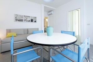 A dining area with a round table, blue chairs, a sofa with a yellow pillow, and a wall-mounted picture.