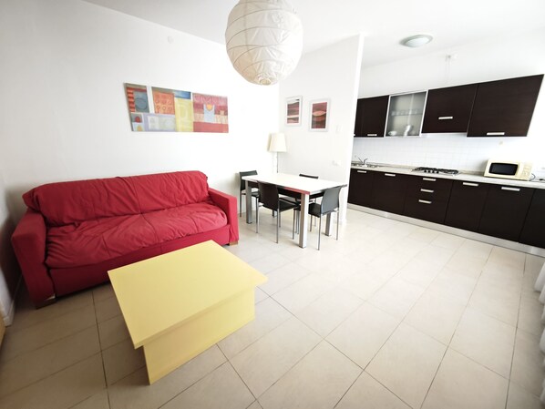 A modern living room with a red sofa, a yellow table, a dining area, and a kitchen with dark cabinets and a microwave.