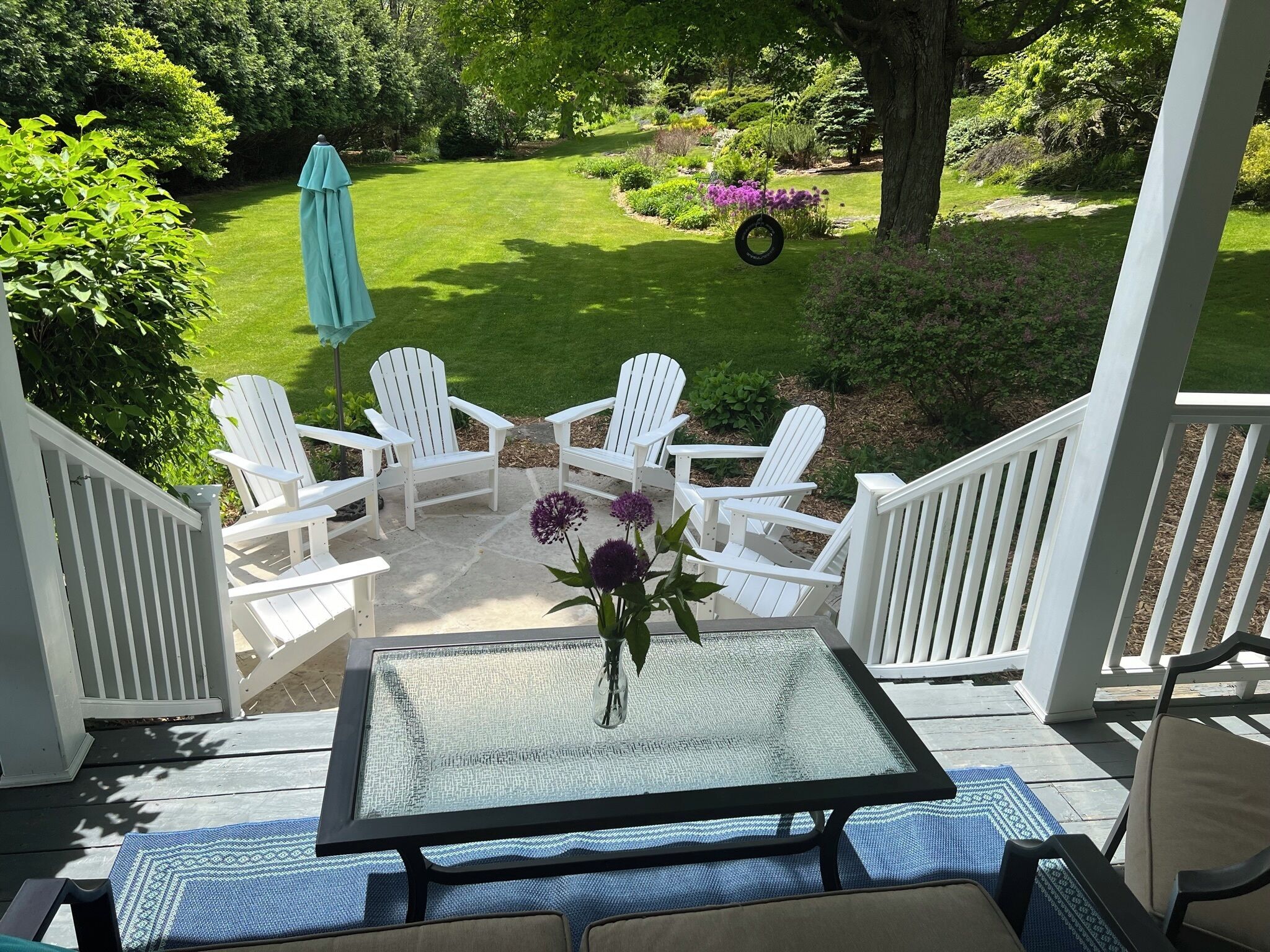 A covered patio with a glass table, white Adirondack chairs, and a view of a well-maintained garden.