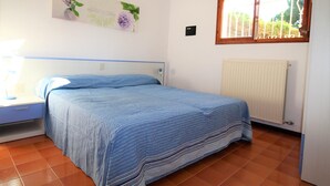 A bedroom with a bed, a nightstand, a wall-mounted lamp, a radiator, and a window with curtains.