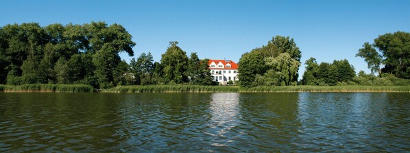 A lakeside view with a white building with a red roof, surrounded by dense greenery. - Family vacation on the lake (Behren-Lübchin)