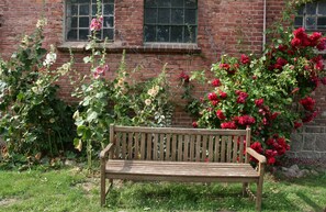 Eine hölzerne Bank in einem Garten mit blühenden Blumen und einer Backsteinmauer im Hintergrund.