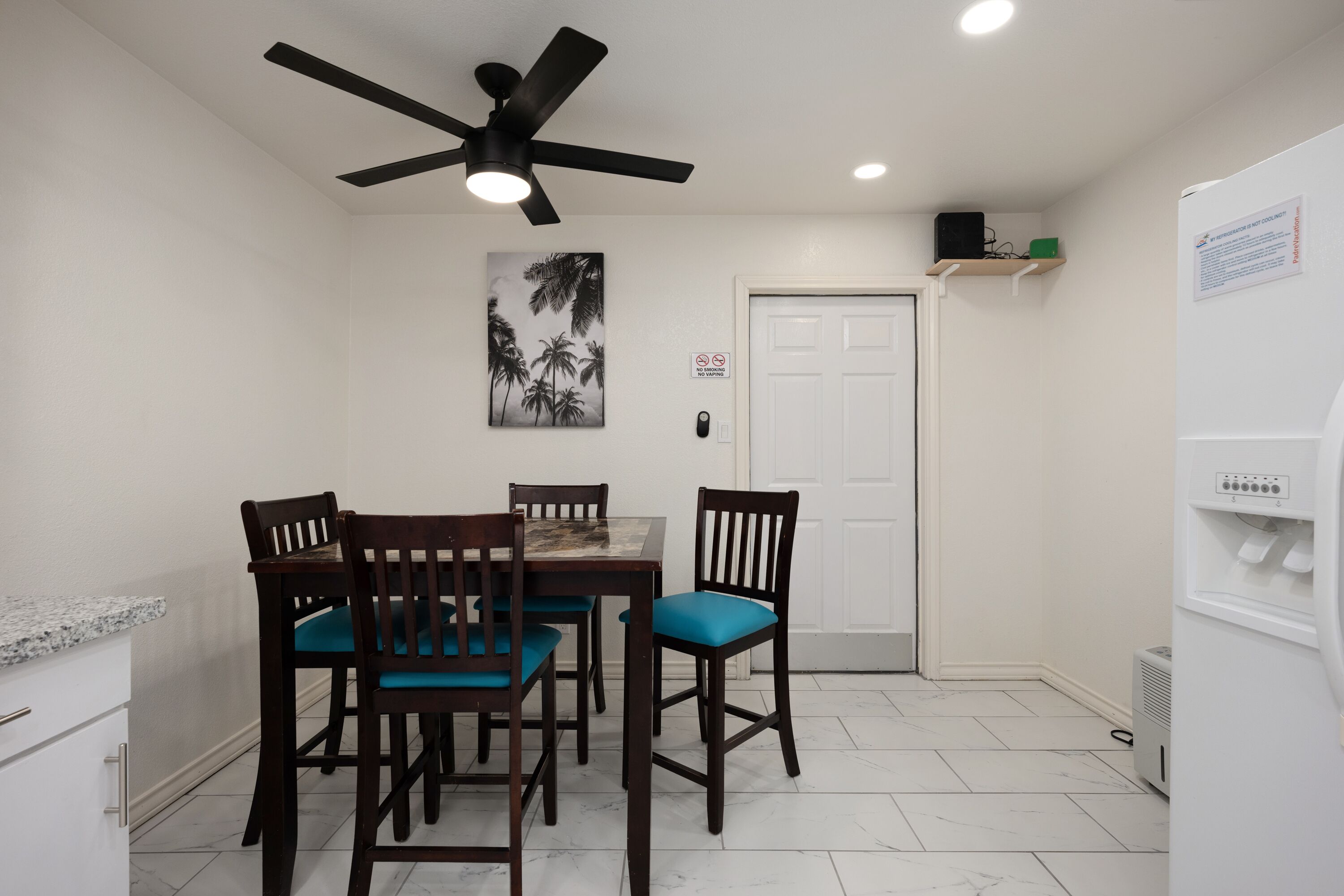 Un comedor con una mesa, sillas, un ventilador de techo, una puerta y un refrigerador.