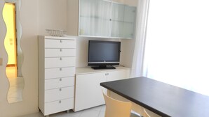 A small, modern kitchen with a white cabinet, a black television, and a dining table with two chairs. - Condominio Billa 8 - Lignano (Lignano Sabbiadoro)