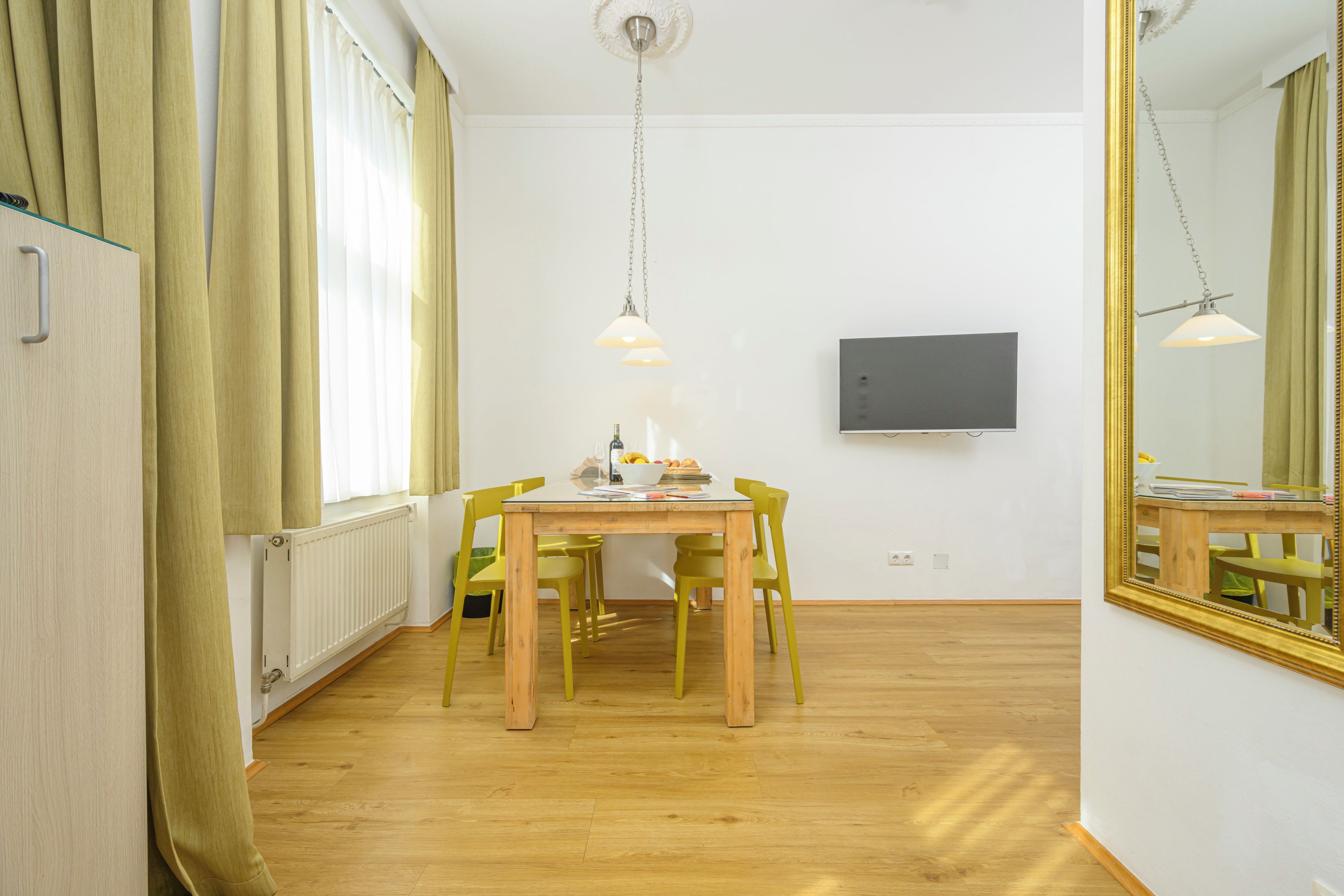 A room with a wooden floor, a dining table with chairs, a television on the wall, and a large mirror.