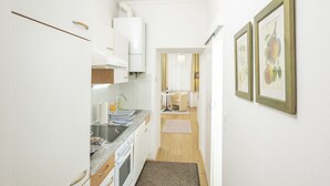 A narrow hallway leading to a kitchen with white cabinets, a stove, and a sink. There is a small table with a chair in the background.
