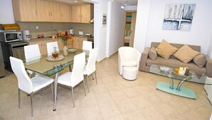 Une cuisine moderne avec des meubles en bois, un coin repas doté d’une table en verre et de chaises blanches, et un salon avec un canapé beige et une table basse en verre.