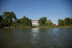 A house by a calm river with a red roof and white walls, surrounded by trees.