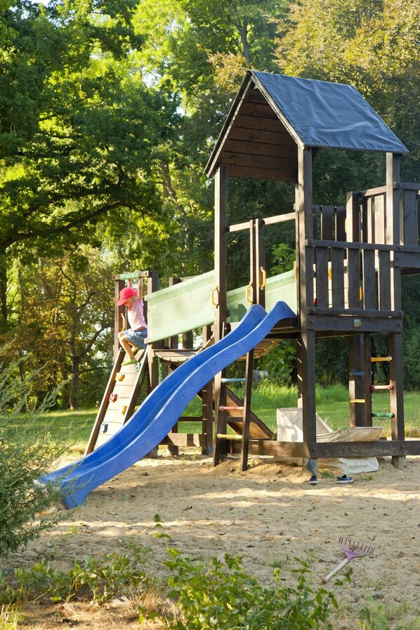 A wooden play structure with a slide and climbing area, surrounded by trees and grass.