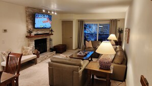 A living room with a stone fireplace, a television mounted on the wall, a sofa, a coffee table, and a lamp. - Main St/Town Lift/Hot Tub/Parking (Park City)