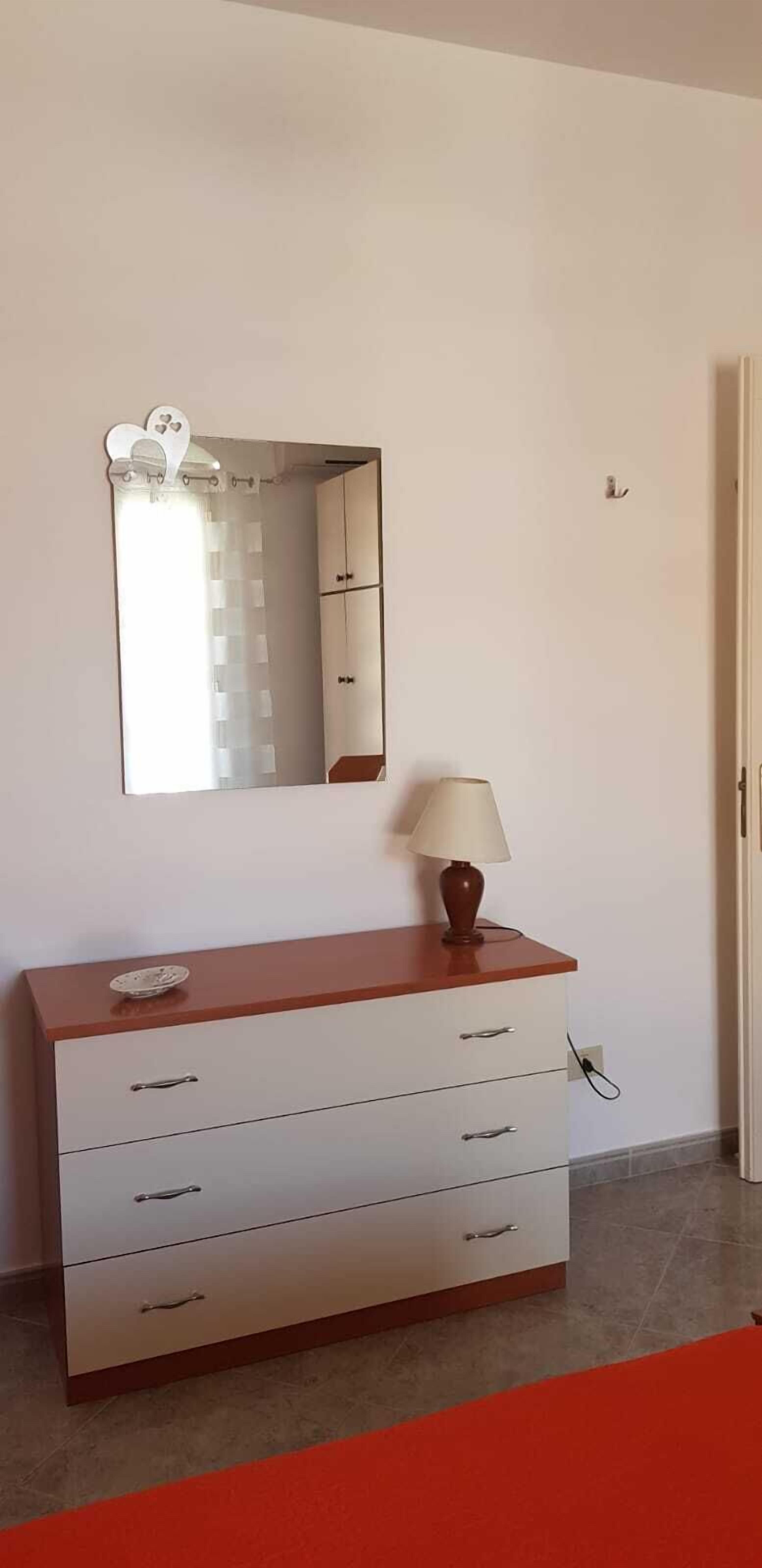 Een witte dressoir met laden, een lamp en een spiegel met een hartvormig frame, plus een rood tapijt.
