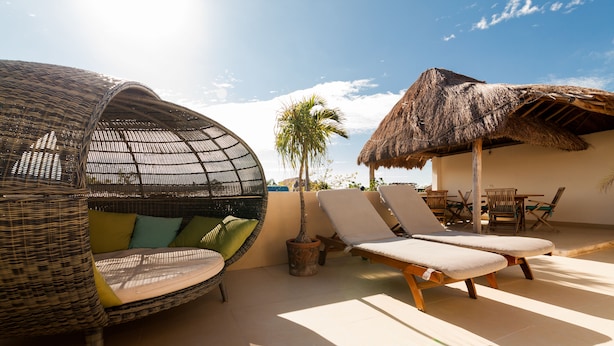 Un área lounge al aire libre con luz solar, mobiliario de mimbre, una estructura de techo de paja y vistas al cielo.