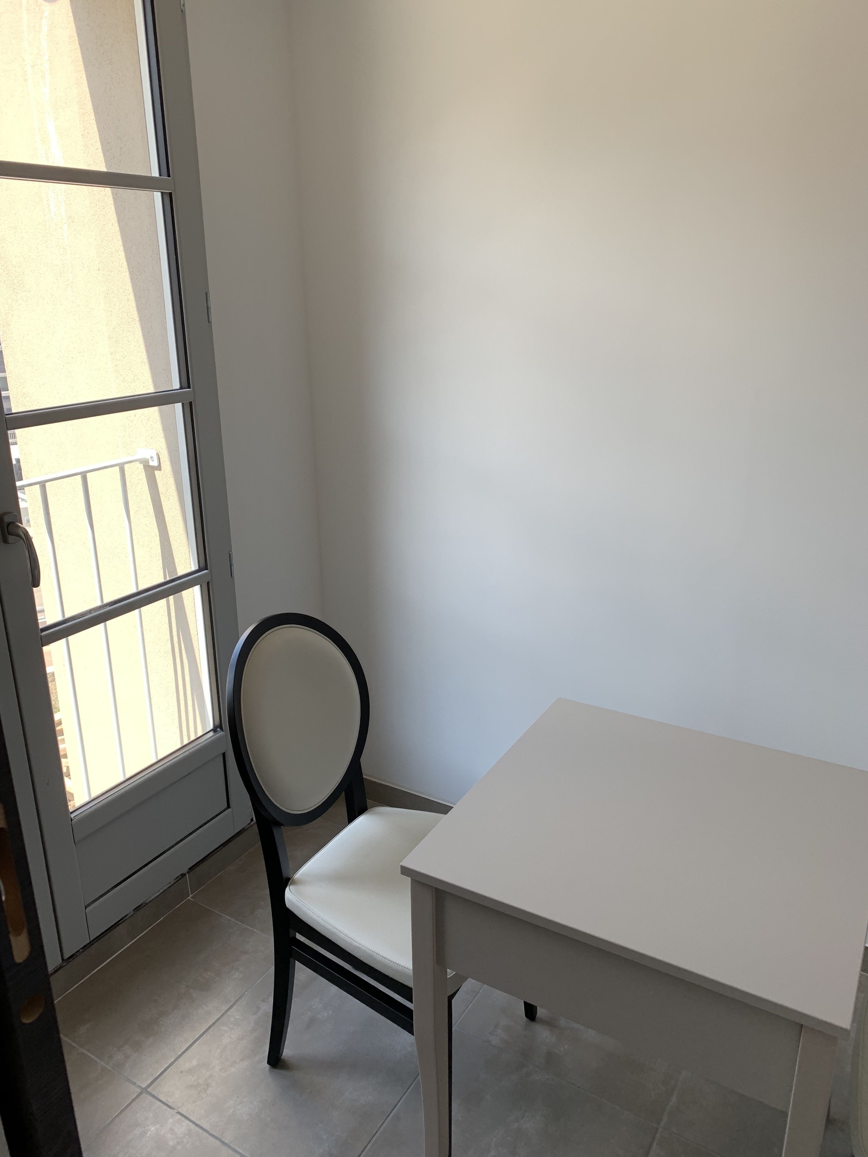A corner room with a white table and a chair, a window with a view of the outside, and a door.