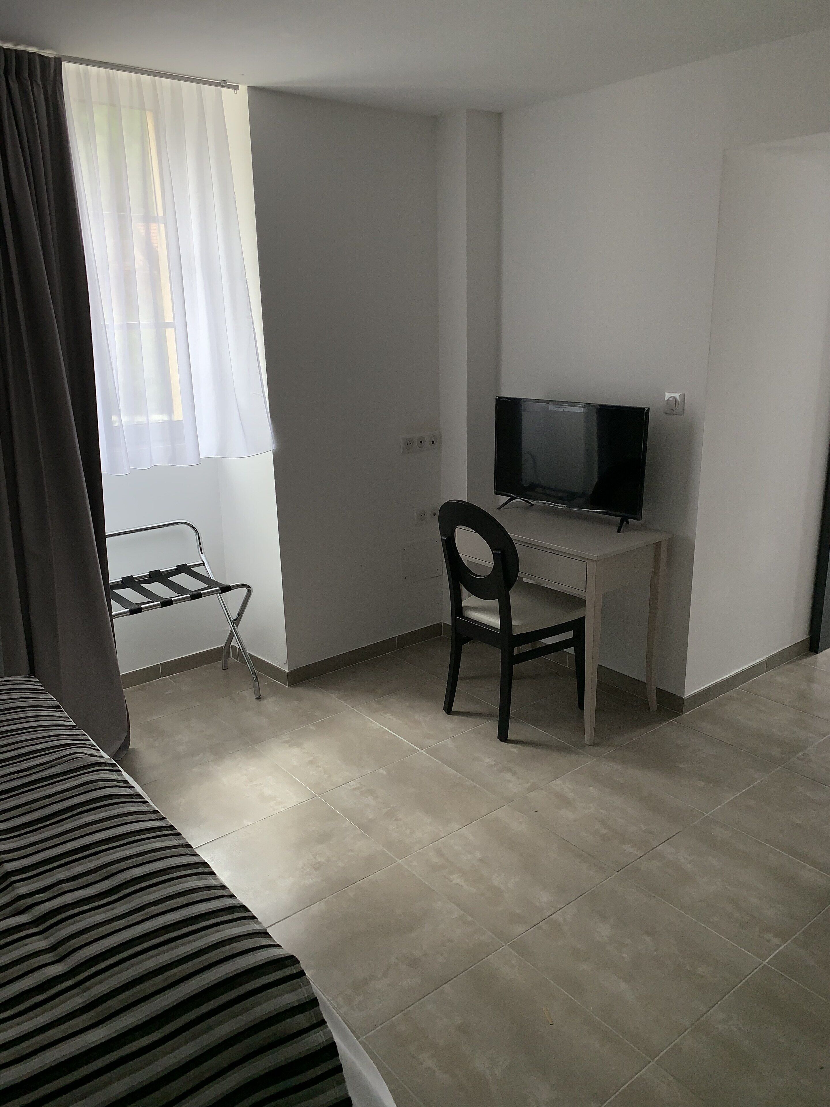 Una habitación de hotel con cama, escritorio con silla, televisor y ventana con cortinas.