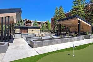 Uma área externa moderna com campo de putting, assentos e um edifício com varanda coberta.