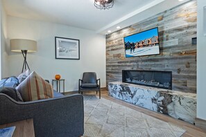 Uma sala de estar moderna com lareira, uma TV de tela plana fixada em uma parede de madeira, um sofá com almofadas estampadas e uma mesinha de cabeceira com abajur.