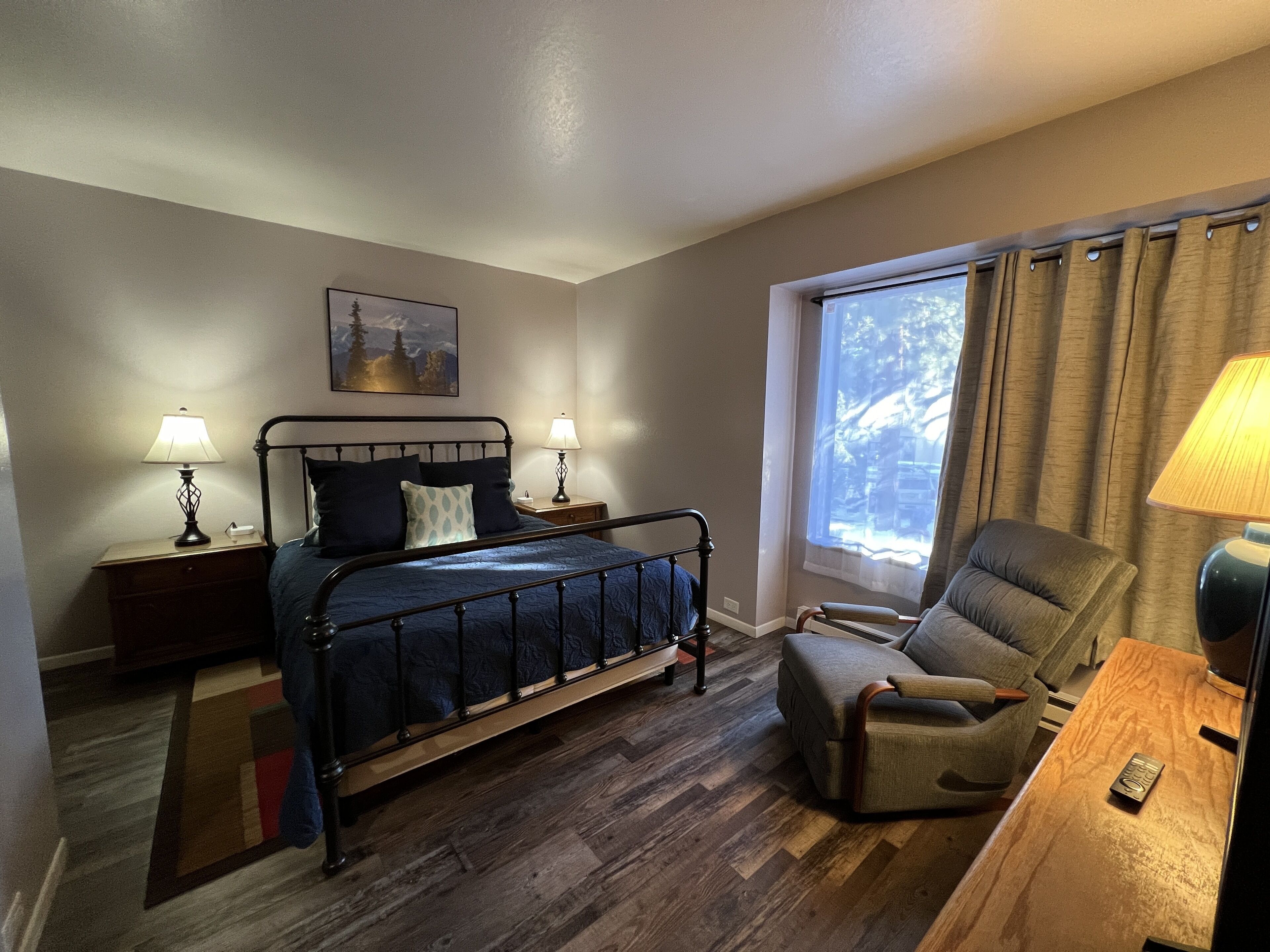 Une chambre à coucher avec un lit, des lampes de chevet, une chaise, une fenêtre avec des rideaux et un tableau au mur.